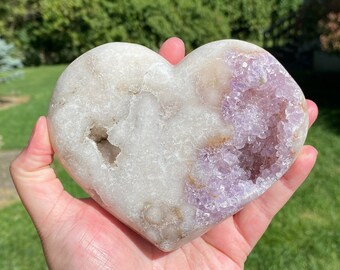 Puede incluir: Un cristal de amatista en forma de corazón con un exterior natural y en bruto y un centro hueco lleno de cristales de amatista más pequeños. El cristal es de color púrpura claro y tiene un aspecto blanco y nublado.