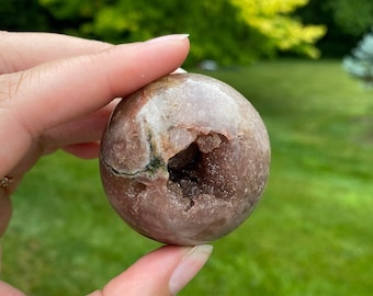 May include: A pink and brown geode sphere with a crystal cave inside. The sphere is smooth and polished, with a natural, organic shape.