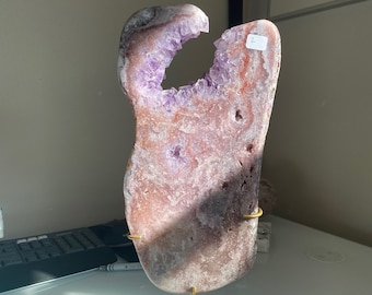 Puede incluir: Una gran geoda de amatista de forma libre con una abertura natural, que muestra cristales púrpuras. La geoda está montada en un soporte de metal dorado.