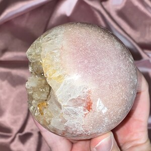 Puede incluir: Una esfera de geoda de &aacute;gata rosa y blanca con una gran abertura que revela un c&uacute;mulo de cristales en su interior. La geoda es lisa y pulida.