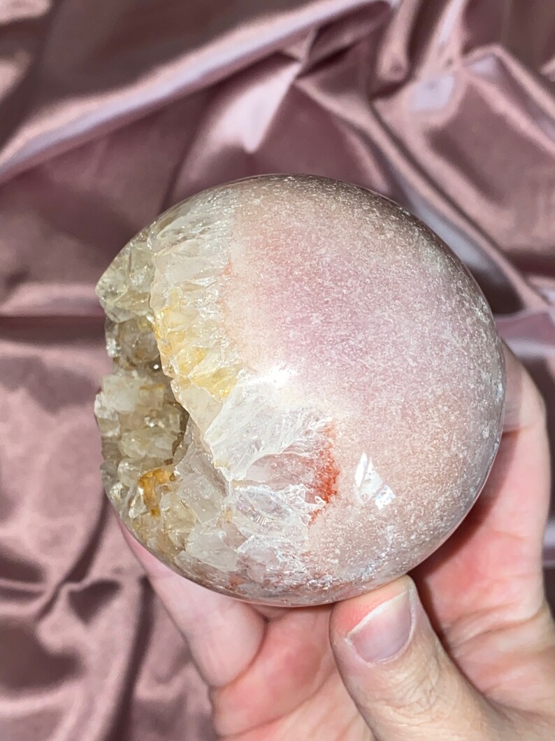 Puede incluir: Una esfera de geoda de &aacute;gata rosa y blanca con una gran abertura que revela un c&uacute;mulo de cristales en su interior. La geoda es lisa y pulida.