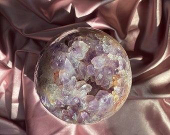 Puede incluir: Una gran esfera de geoda de amatista con una cáscara exterior transparente y un racimo de cristales morados en su interior. La geoda está sentada sobre una tela de satén rosa.
