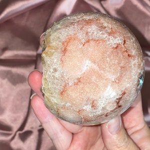 Puede incluir: Una gran esfera de cristal redonda y transparente con inclusiones rojas y blancas. La esfera es lisa y pulida, y tiene una apariencia transl&uacute;cida.