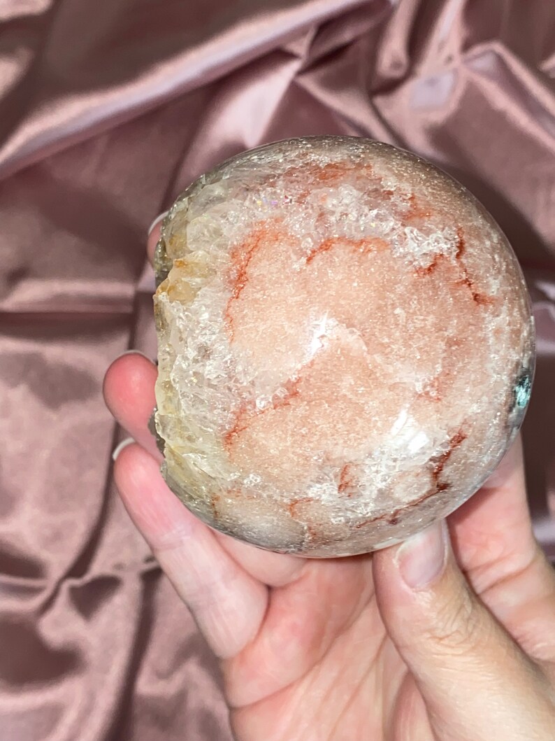 Puede incluir: Una gran esfera de cristal redonda y transparente con inclusiones rojas y blancas. La esfera es lisa y pulida, y tiene una apariencia transl&uacute;cida.