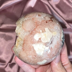 Puede incluir: Una esfera de geoda transl&uacute;cida rosa, blanca y marr&oacute;n con una superficie rugosa e irregular. La geoda tiene un aspecto natural y org&aacute;nico.