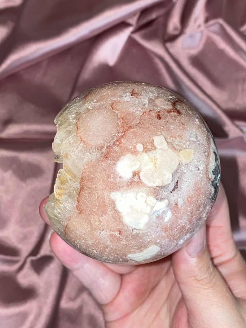 Puede incluir: Una esfera de geoda transl&uacute;cida rosa, blanca y marr&oacute;n con una superficie rugosa e irregular. La geoda tiene un aspecto natural y org&aacute;nico.