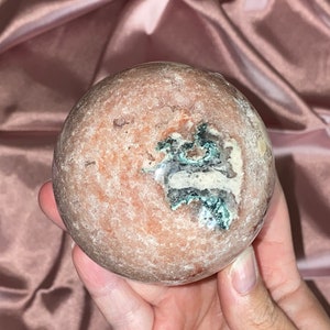 Puede incluir: Una esfera pulida de &aacute;gata rosa y blanca con una inclusi&oacute;n verde y blanca. La esfera es lisa y tiene una textura natural y org&aacute;nica.
