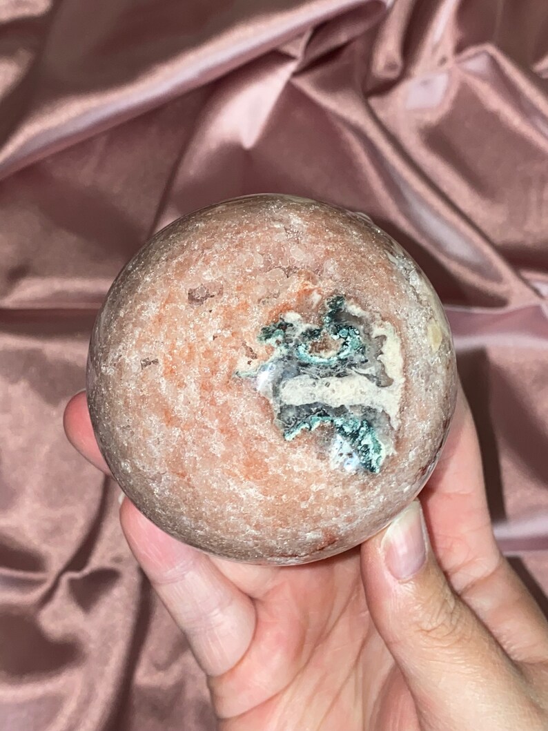 Puede incluir: Una esfera pulida de &aacute;gata rosa y blanca con una inclusi&oacute;n verde y blanca. La esfera es lisa y tiene una textura natural y org&aacute;nica.