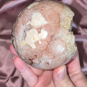 Puede incluir: Una gran geoda redonda de color rosa y blanco con una abertura natural. La geoda tiene una superficie rugosa y texturizada y est&aacute; llena de cristales blancos.