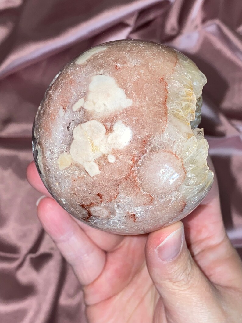 Puede incluir: Una gran geoda redonda de color rosa y blanco con una abertura natural. La geoda tiene una superficie rugosa y texturizada y est&aacute; llena de cristales blancos.