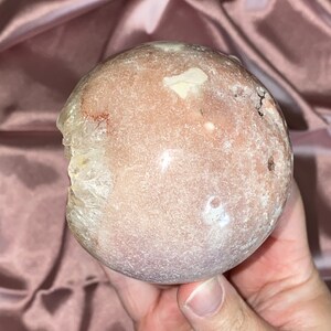 Puede incluir: Una esfera de geoda rosa y blanca con una superficie rugosa y texturizada. La geoda tiene una abertura natural que revela un cristal claro en el interior.