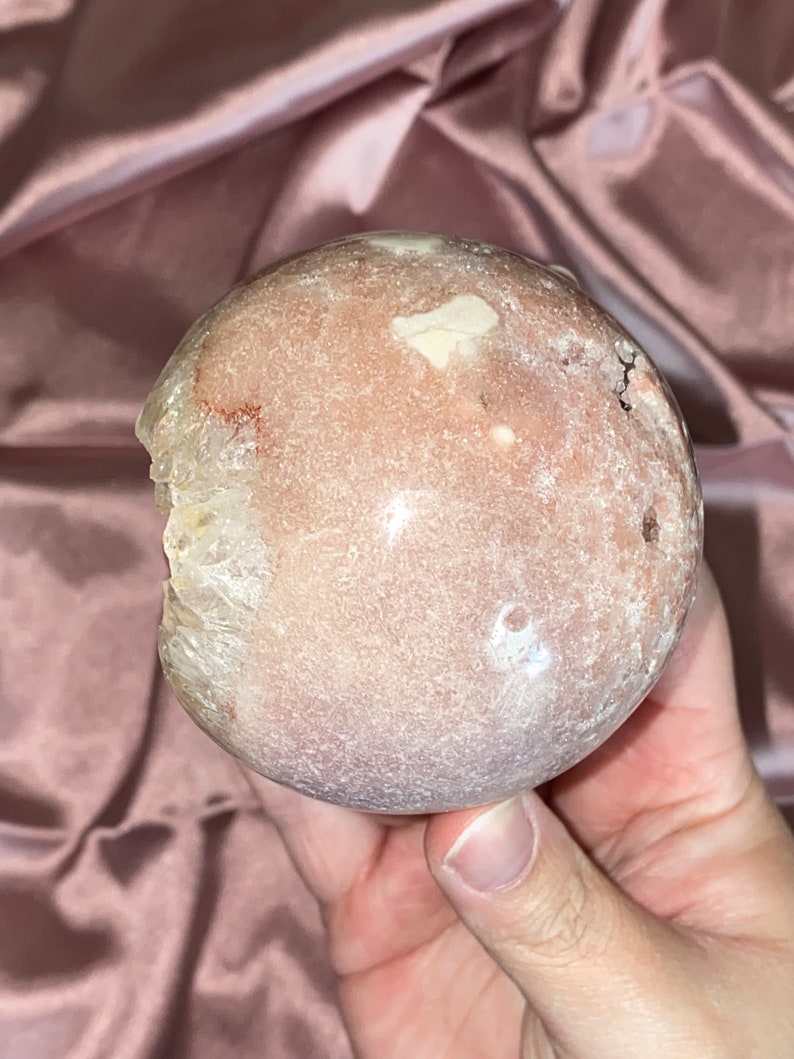 Puede incluir: Una esfera de geoda rosa y blanca con una superficie rugosa y texturizada. La geoda tiene una abertura natural que revela un cristal claro en el interior.