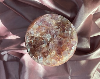 Puede incluir: Una gran geoda de amatista con un exterior natural y bruto y un interior hueco lleno de racimos de cristales de amatista más pequeños. La geoda es de color púrpura claro y tiene una superficie lisa y pulida.