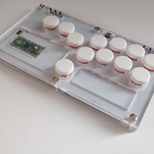 May include: Clear acrylic arcade controller with white buttons and red accents. The controller has a rectangular shape with a small circuit board and gold-colored screws. The buttons are arranged in two rows.