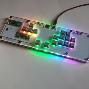 Puede incluir: Un teclado personalizado con una base de acrílico transparente y teclas grises. El teclado cuenta con iluminación LED multicolor a lo largo de los bordes y debajo de las teclas. Un cable rojo está conectado.