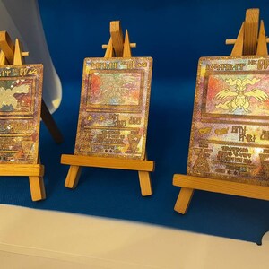 以下が含まれることがあります： 小さな木製のイーゼルに飾られた、ピカチュウのキャラクターが描かれたホログラフィックポケモンカード3枚。カードはゴールド、シルバー、ブロンズで、虹色の輝きを放っています。