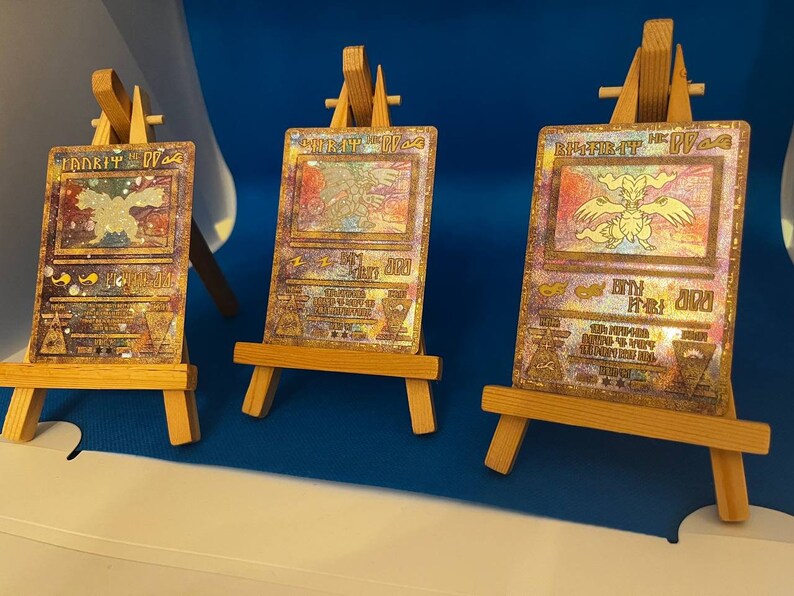 以下が含まれることがあります： 小さな木製のイーゼルに飾られた、ピカチュウのキャラクターが描かれたホログラフィックポケモンカード3枚。カードはゴールド、シルバー、ブロンズで、虹色の輝きを放っています。