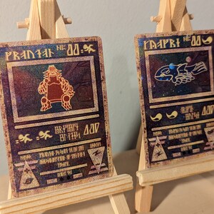 May include: Two vintage-style trading cards with holographic foil accents. The card on the left features a red monster with the text "PRANIAL NO. 00-.", while the card on the right features a blue creature with the text "MAPRI NO. 00-."