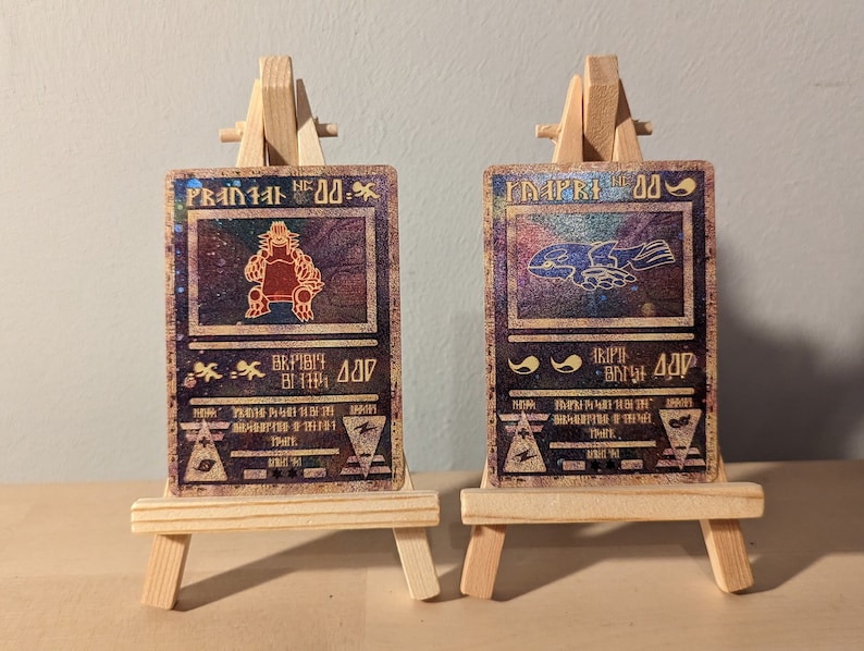 May include: Two fantasy trading cards on wooden easels. The cards feature a red and brown creature with a crown and a blue and purple creature with wings. The cards have a gold and black background with a holographic effect.