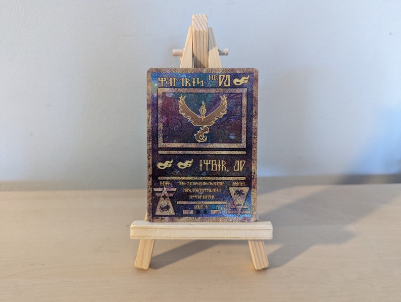 Pode incluir: Um cart&atilde;o Pokemon dourado e azul com uma imagem hologr&aacute;fica do Pokemon Moltres. O cart&atilde;o est&aacute; em um pequeno cavalete de madeira.