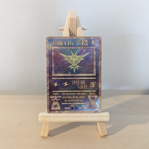 Pode incluir: Um cart&atilde;o Pok&eacute;mon dourado e azul com acabamento hologr&aacute;fico. O cart&atilde;o apresenta um raio amarelo e o texto "Pikachu" em japon&ecirc;s. O cart&atilde;o &eacute; exibido em um cavalete de madeira.