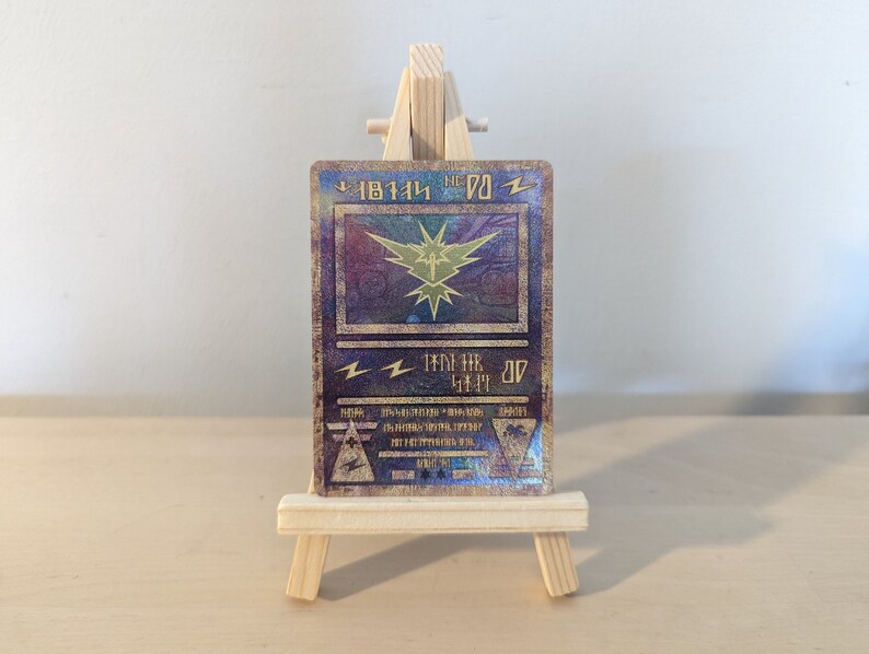 Pode incluir: Um cart&atilde;o Pok&eacute;mon dourado e azul com acabamento hologr&aacute;fico. O cart&atilde;o apresenta um raio amarelo e o texto "Pikachu" em japon&ecirc;s. O cart&atilde;o &eacute; exibido em um cavalete de madeira.