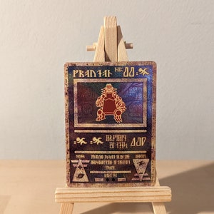 May include: A gold and red holographic trading card with a picture of a red monster with a crown on its head. The card is titled "PRANIAL DD-" and has a black and white background with a gold border.