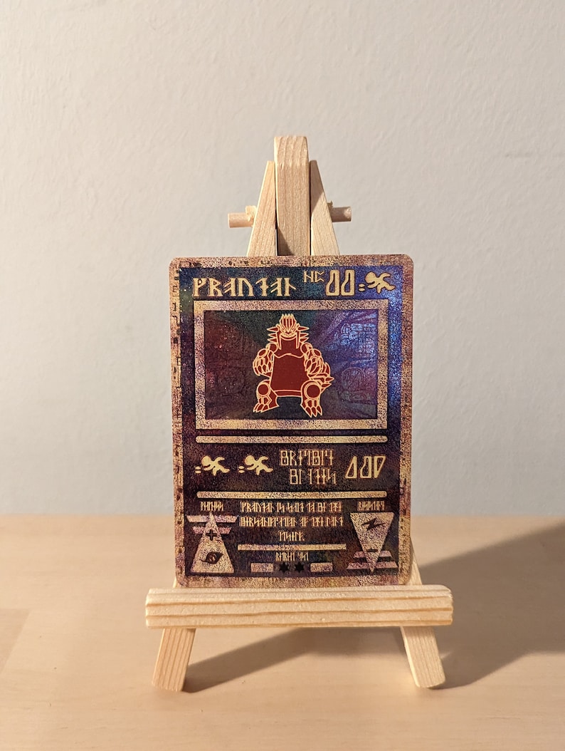 May include: A gold and red holographic trading card with a picture of a red monster with a crown on its head. The card is titled "PRANIAL DD-" and has a black and white background with a gold border.