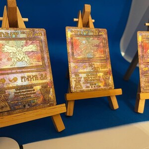 以下が含まれることがあります： 「ダーク・マジシャン・ガール」カードが描かれた金色の遊戯王トレーディングカードが3枚。カードは小さな木製イーゼルに飾られています。