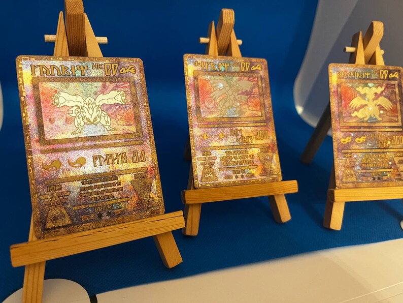 以下が含まれることがあります： 「ダーク・マジシャン・ガール」カードが描かれた金色の遊戯王トレーディングカードが3枚。カードは小さな木製イーゼルに飾られています。