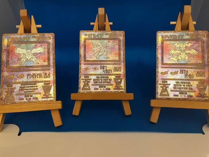 以下が含まれることがあります： 虹色の効果が施された白い背景に、キャラクター「レックウザ」が描かれた金色のポケモンカードが3枚。カードは小さな木製イーゼルに飾られています。