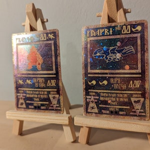 May include: Two holographic trading cards with a gold border and a black background. The cards feature illustrations of mythical creatures and text in a fantasy font. The cards are displayed on a wooden easel.