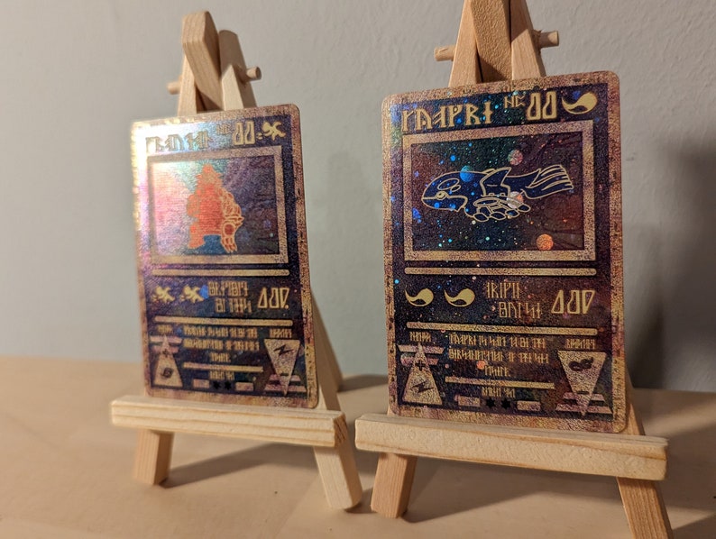 May include: Two holographic trading cards with a gold border and a black background. The cards feature illustrations of mythical creatures and text in a fantasy font. The cards are displayed on a wooden easel.