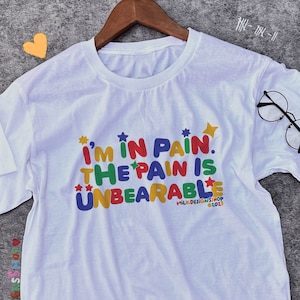 May include: White t-shirt with a colourful graphic that says "I'm in pain. The pain is unbearable."  The graphic is in a playful font with stars around the text.