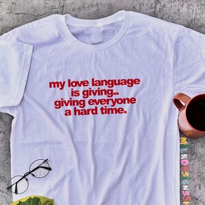 May include: White t-shirt with the red text "my love language is giving.. giving everyone a hard time." The shirt is next to a pair of round glasses, a leaf, and a copper-coloured mug filled with a dark liquid.