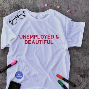 May include: White t-shirt with the text "UNEMPLOYED & BEAUTIFUL" printed in red. The shirt is lying flat on a gray surface with a pair of glasses, a pink marker, a blue sticker with the text "MILK DESIGNS", a black phone, and orange and green markers.