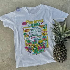 May include: White t-shirt featuring a colourful graphic of pineapples playing musical instruments. The graphic includes the text "Pineapple Jam!" and "Recipe: Pick fresh, juicy pineapples. Add a full set of musical instruments. Shake! Rattle and Roll!"