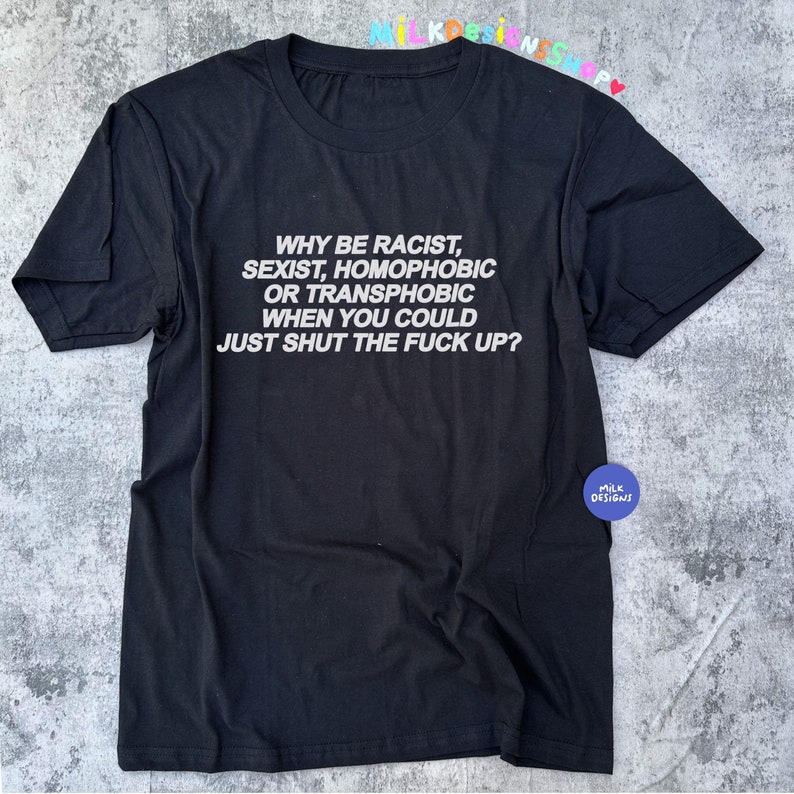 May include: Black t-shirt with white text: "WHY BE RACIST, SEXIST, HOMOPHOBIC OR TRANSPHOBIC WHEN YOU COULD JUST SHUT THE FUCK UP?". The shirt has short sleeves and a round neckline. A small circular logo is visible.