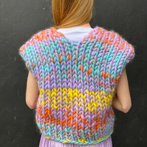May include: A sleeveless knitted vest with a colourful striped pattern in shades of purple, orange, yellow, and blue. The vest is made with chunky yarn and has a relaxed fit.