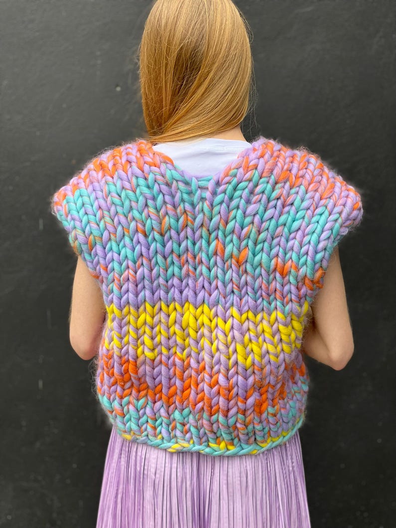 May include: A sleeveless knitted vest with a colourful striped pattern in shades of purple, orange, yellow, and blue. The vest is made with chunky yarn and has a relaxed fit.