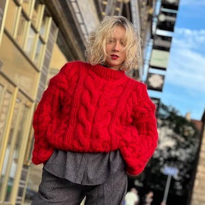 May include: A red chunky knit cable sweater with a round neckline. The sweater is oversized and has a relaxed fit.