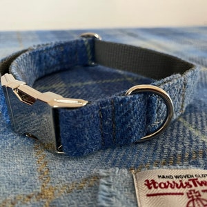 May include: A blue plaid dog collar with a silver metal buckle and a D-ring. The collar is made of Harris Tweed fabric and has a gray interior lining. The collar is displayed on a matching plaid fabric.