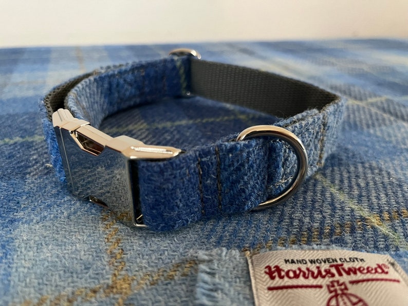 May include: A blue plaid dog collar with a silver metal buckle and a D-ring. The collar is made of Harris Tweed fabric and has a gray interior lining. The collar is displayed on a matching plaid fabric.