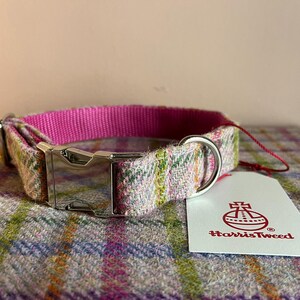 May include: A dog collar with a pink nylon inner lining and a Harris Tweed outer layer in a plaid pattern. The collar features a silver metal buckle and a matching D-ring for leash attachment. A white tag with the Harris Tweed logo is attached.