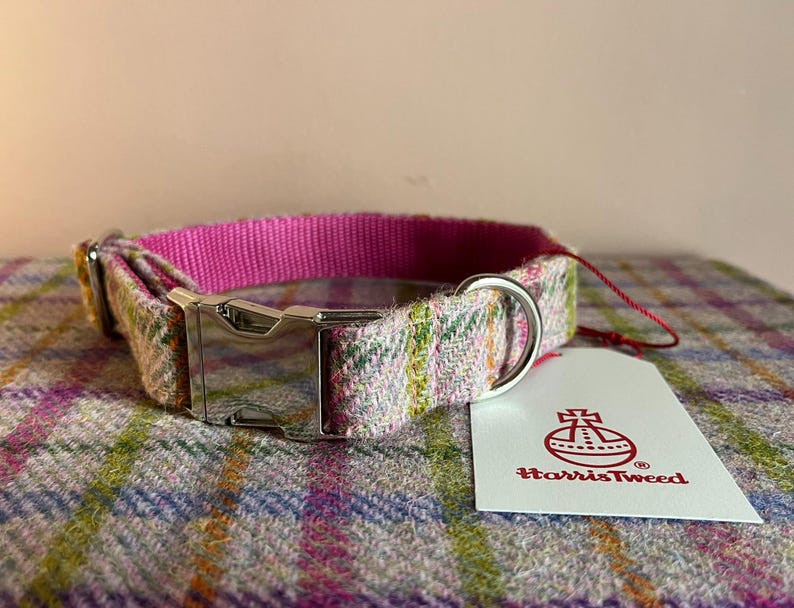 May include: A dog collar with a pink nylon inner lining and a Harris Tweed outer layer in a plaid pattern. The collar features a silver metal buckle and a matching D-ring for leash attachment. A white tag with the Harris Tweed logo is attached.