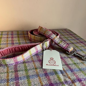 May include: A dog leash made of Harris Tweed fabric, featuring a plaid pattern in pink, green, and yellow hues. The leash has a pink interior and a silver metal clasp. A white tag with the Harris Tweed logo is attached.