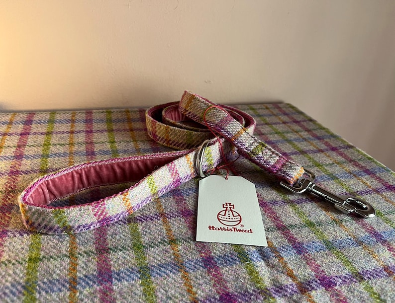 May include: A dog leash made of Harris Tweed fabric, featuring a plaid pattern in pink, green, and yellow hues. The leash has a pink interior and a silver metal clasp. A white tag with the Harris Tweed logo is attached.