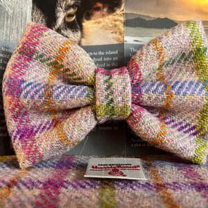 May include: A colorful Harris Tweed bow tie with a plaid pattern in pink, purple, green, and orange. The bow tie is handmade and features a small tag that reads "Harris Tweed". The bow tie is approximately 5 inches wide.