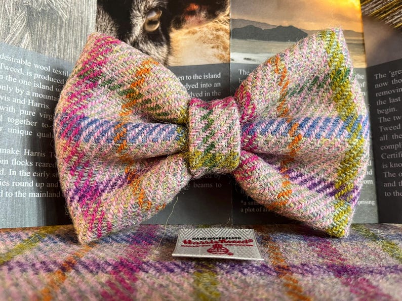 May include: A colorful Harris Tweed bow tie with a plaid pattern in pink, purple, green, and orange. The bow tie is handmade and features a small tag that reads "Harris Tweed". The bow tie is approximately 5 inches wide.