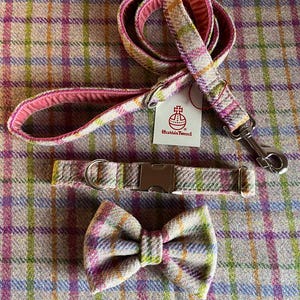May include: A dog leash, collar, and bow tie set. The set features a plaid pattern in pink, green, yellow, and blue. The leash has a pink interior and a metal clasp. The collar has a metal buckle. The bow tie is pre-tied.